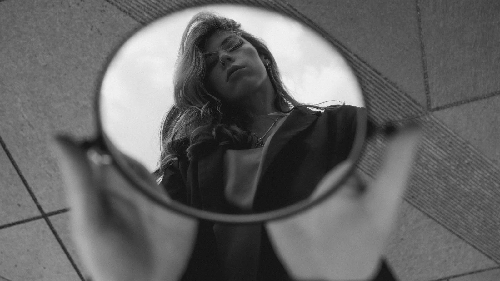 A black-and-white photograph of a woman reflected in a small round mirror held up from the pavement.