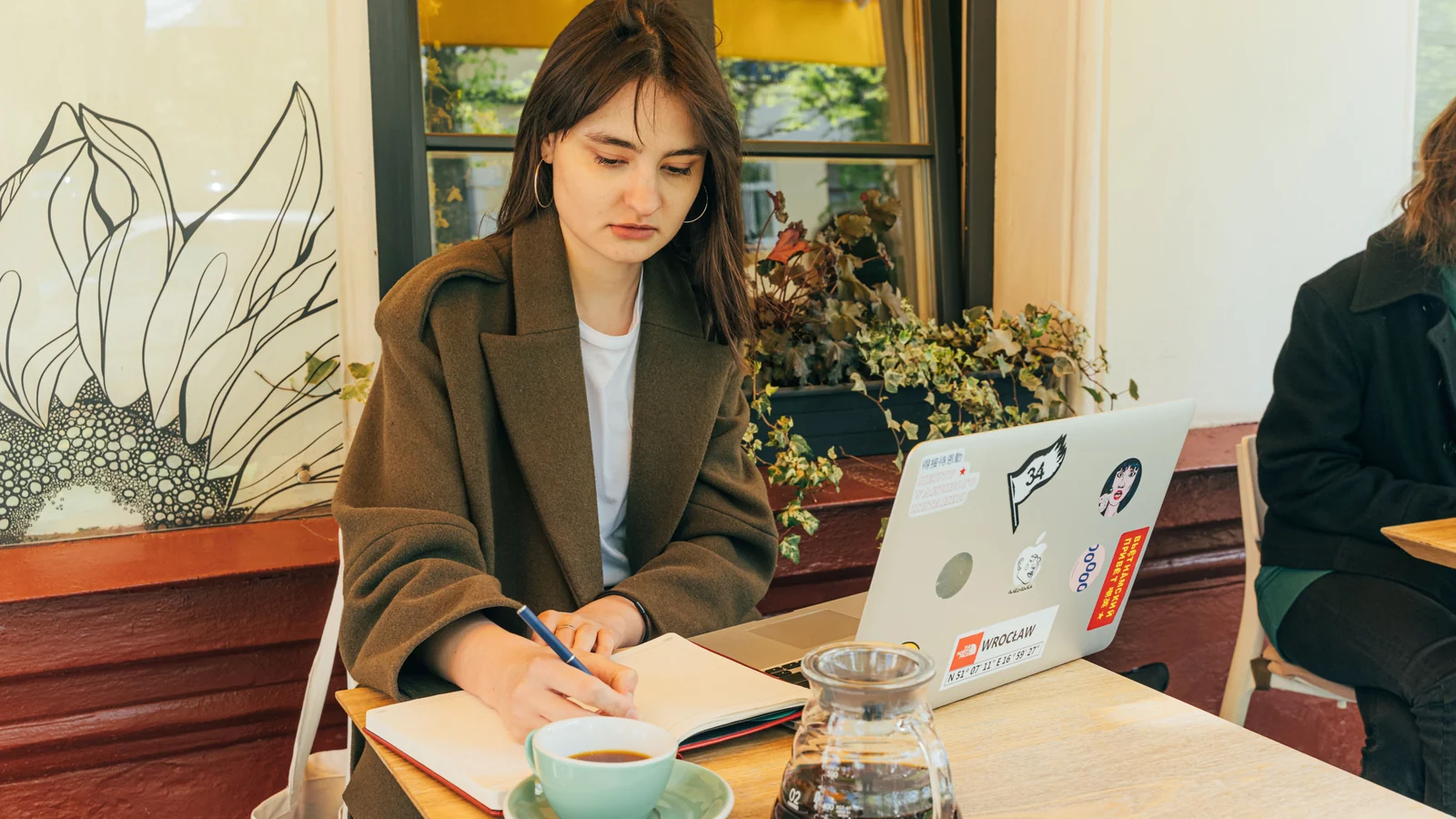 A woman writing in a notebook at an outdoor café table, a laptop and a cup of coffee beside her.