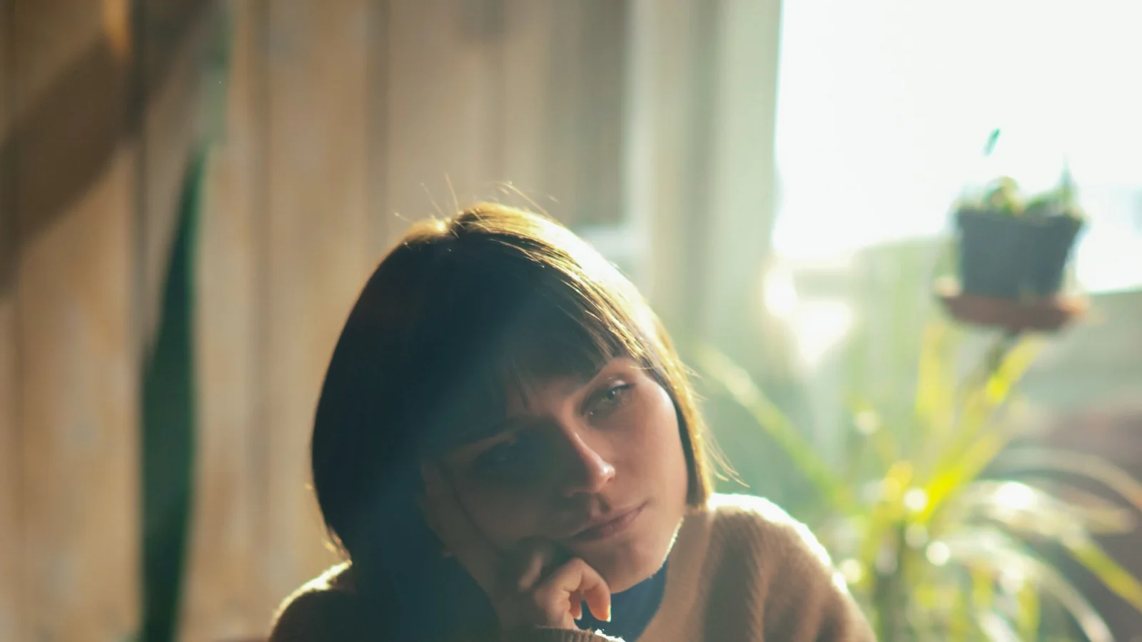 A woman resting her chin on her hand at a laptop, lit by late afternoon light.