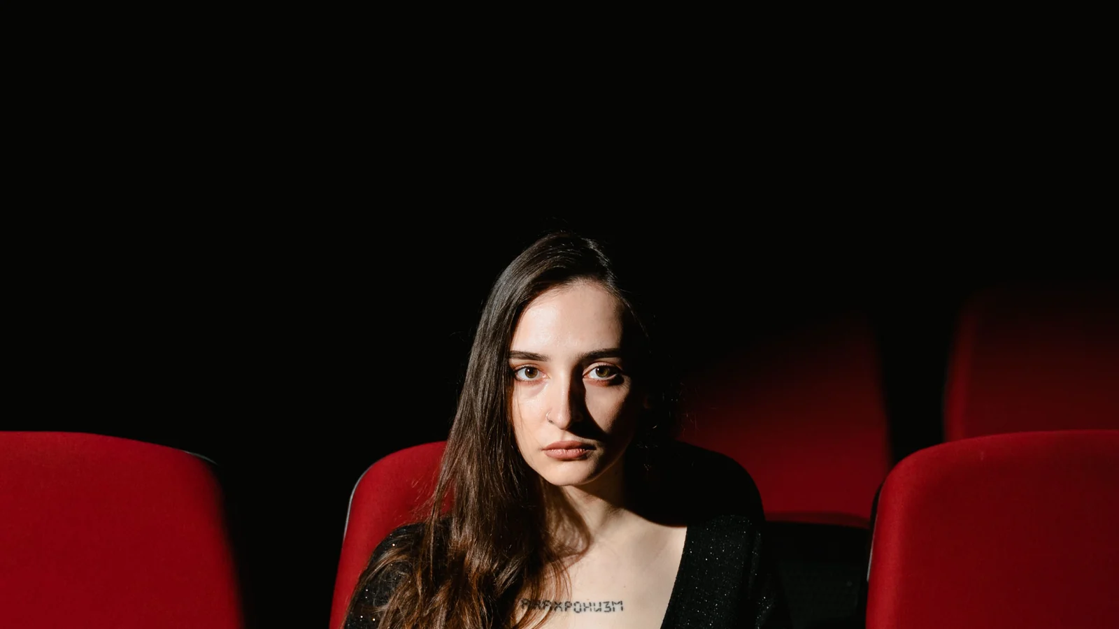 A woman sitting alone in a row of empty red cinema seats, lit by a single narrow beam.