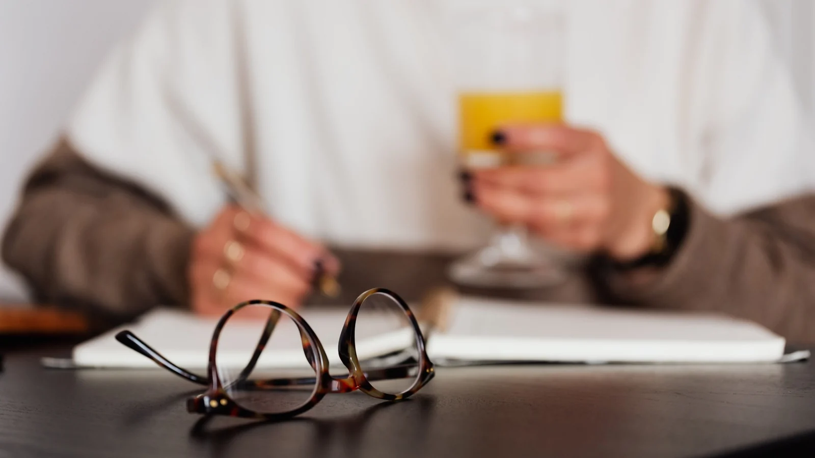A pair of tortoiseshell glasses on a dark table, someone writing in an open notebook behind them.