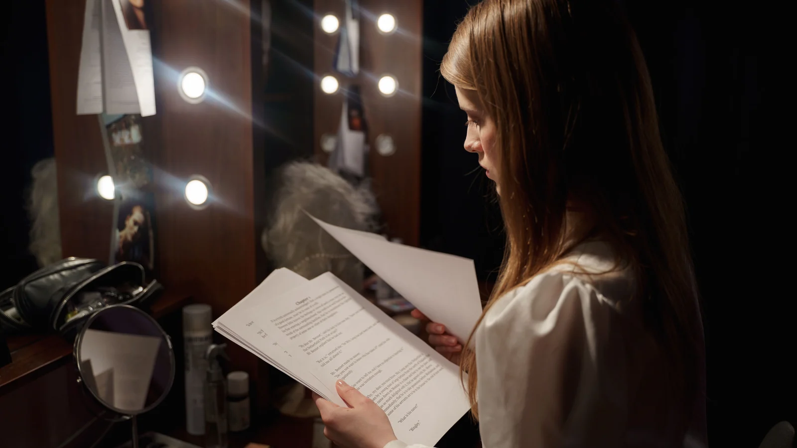 A woman in a dressing room reading from a script, mirror bulbs lit behind her.