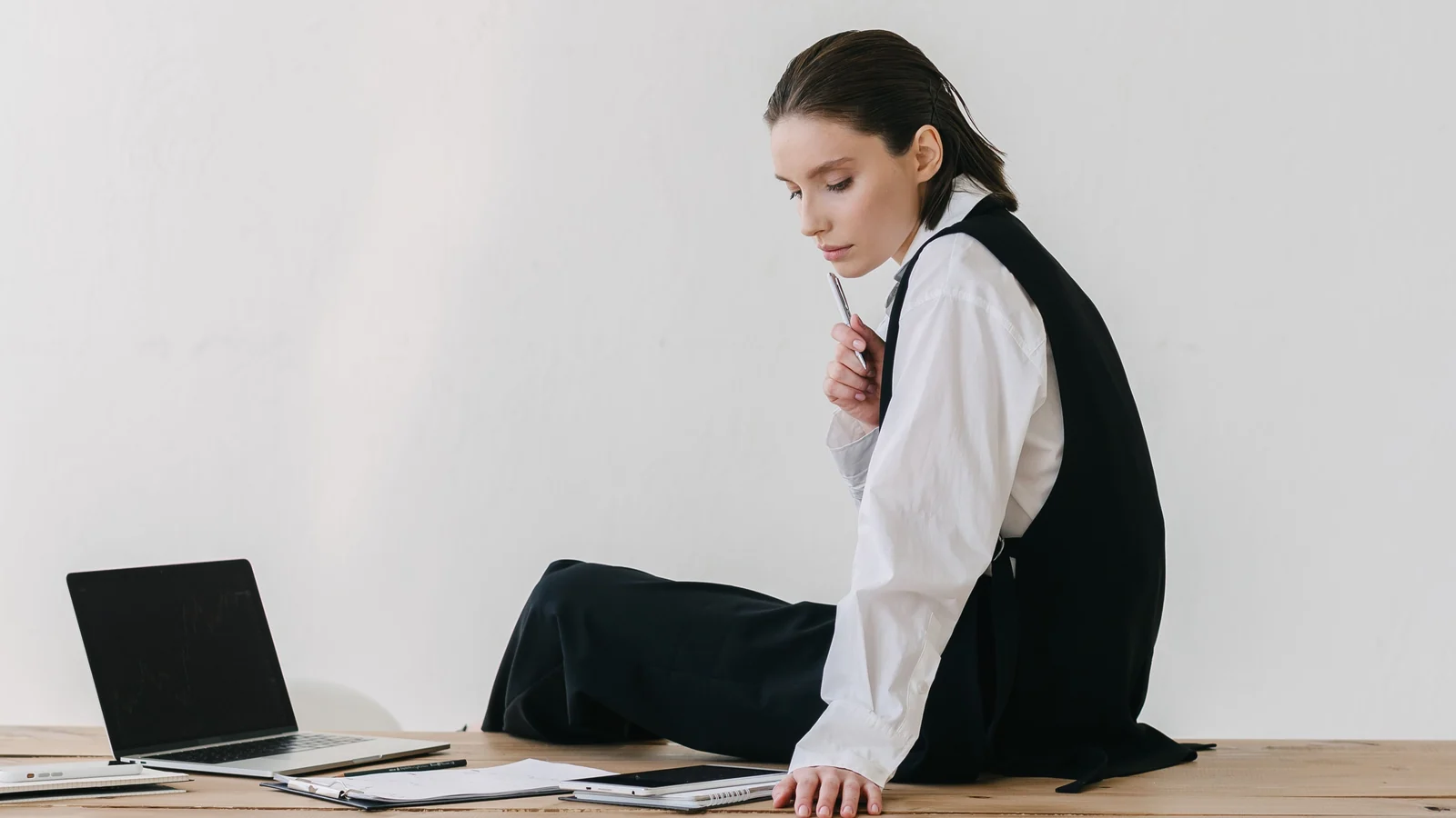 A woman sitting on a wooden desk beside a laptop and notebooks, holding a pen to her chin and looking down.