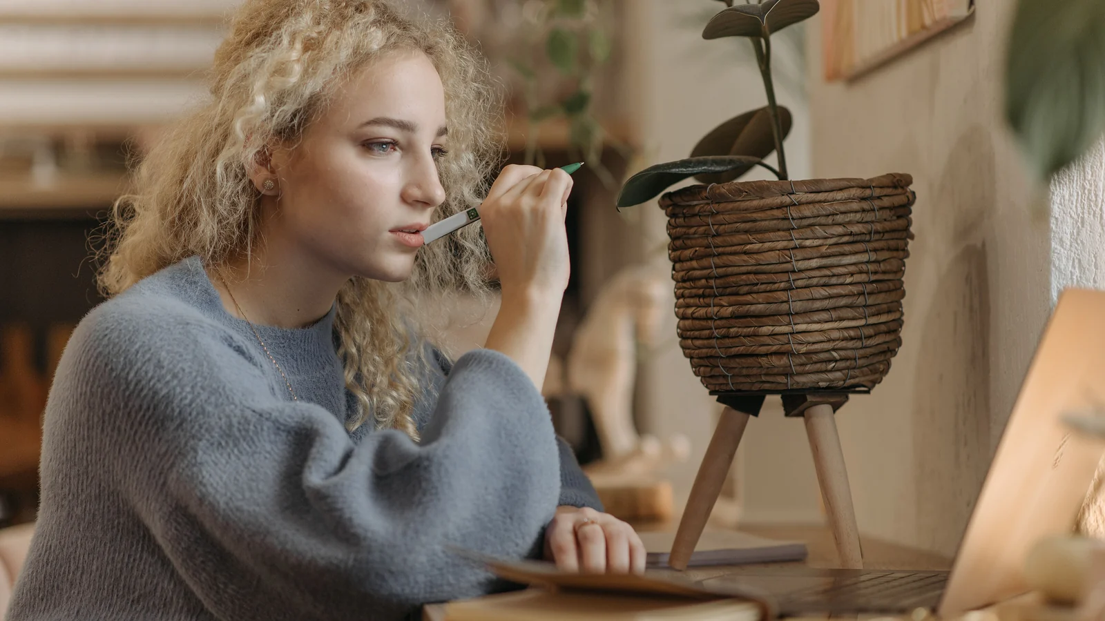 A woman at a desk holding a pen to her lips, looking at a laptop, a plant and warm lamplight behind her.